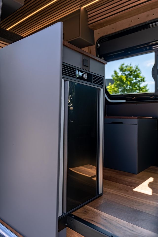 geschlossener Kühlschrank. Eingebaut in ein graues Küchenmöbel. Der Boden ist aus Parket. Wie der Rest des Ausbaus von Cocoon Vans ist der Boden aus amerikanischen Nussbaum. Die Beleuchtunt an der Decke ist eingeschalten.