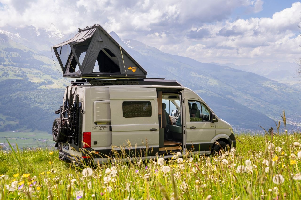 MAN 4x4 Aussenansicht mit geöffnetem Dachzelt, DSRT Heckträger für Velos und einem Reserveradhalter. Das Fahrzeug steht in einer grünen Wiese. Der Himmer ist bewölkt.