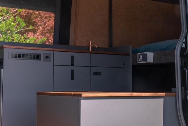 Blick auf die Küche im Camper Van. Das Hubbett ist herunter gelassen. Die Sonne scheint auf die Oberflächen aus amerikanischen Nussbau.